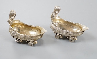 A pair of late 19th/early 20th century Hanau white metal salts, modelled as boat's with wheels and putti surmounts, maker Stork & Sinsheimer, length 81mm, 195 grams.
